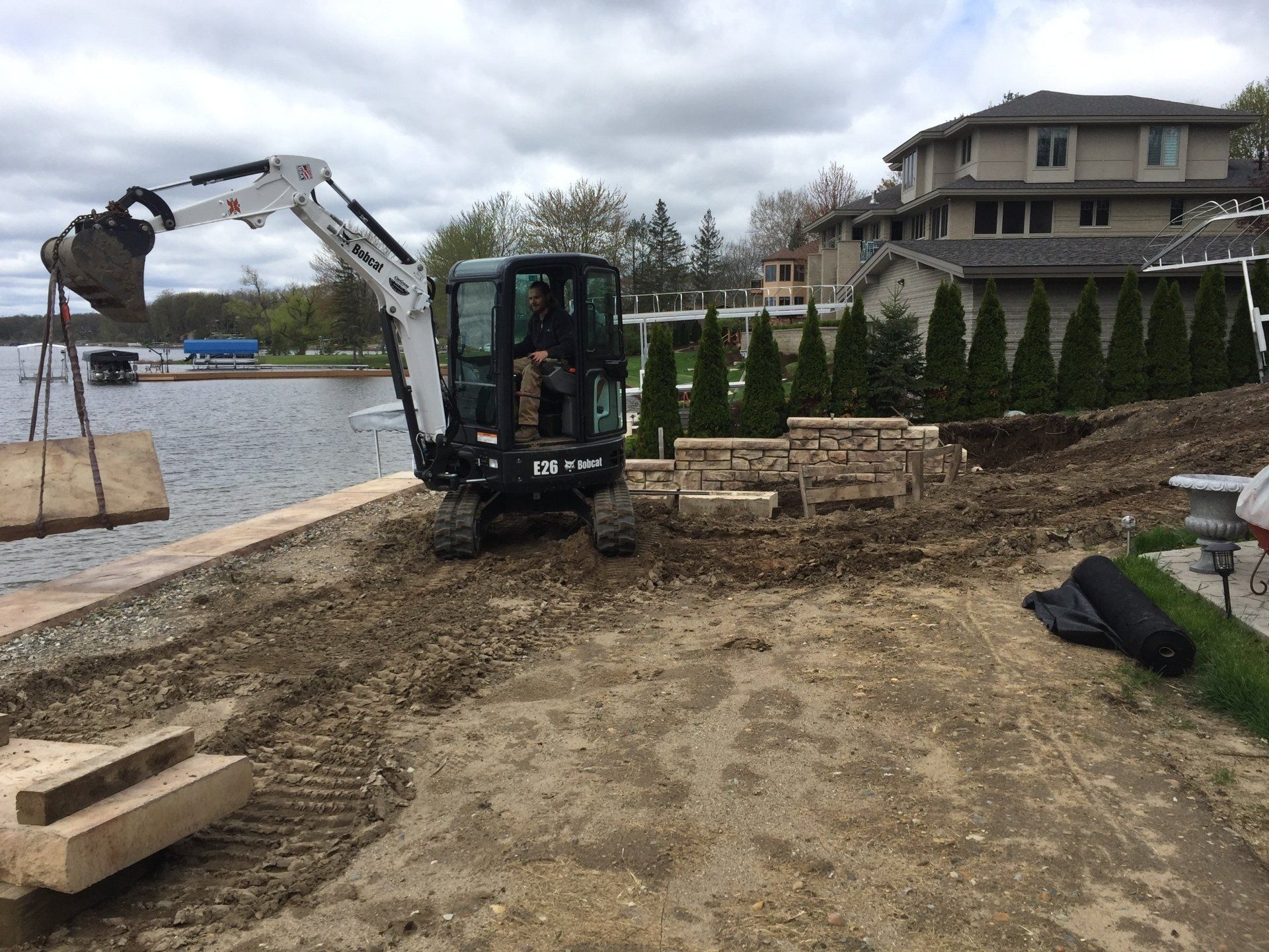 A man is driving an excavator on a dirt road next to a body of water.
