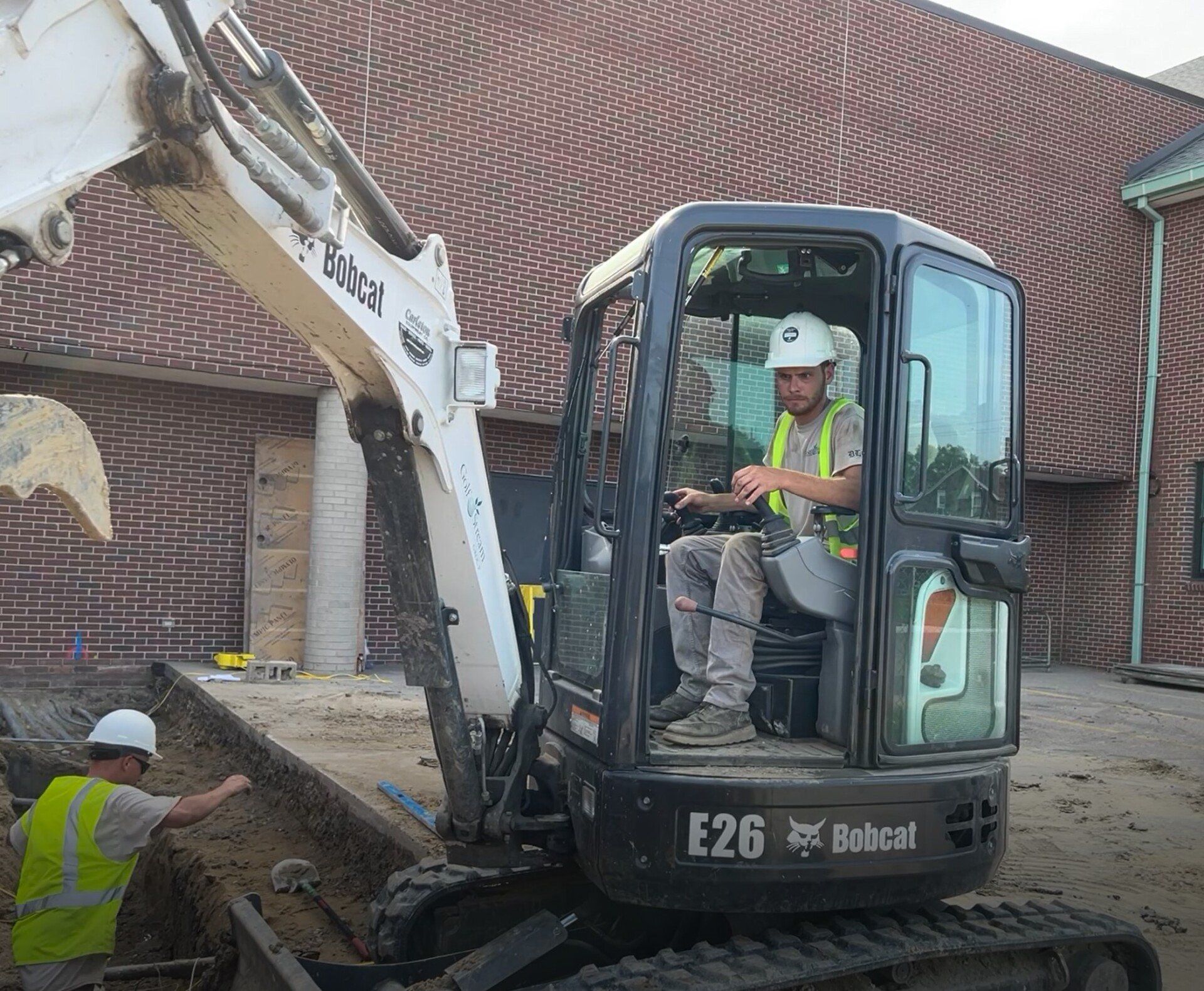 A man is driving a bobcat excavator in front of a brick building.