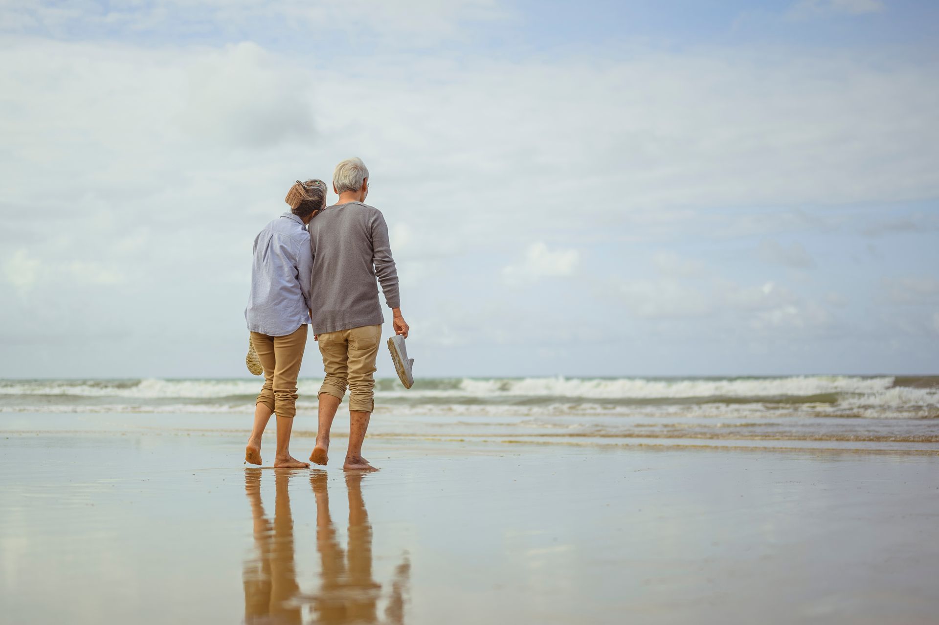 Elderly couple walking barefoot on a wet beach, looking toward the ocean under a cloudy sky