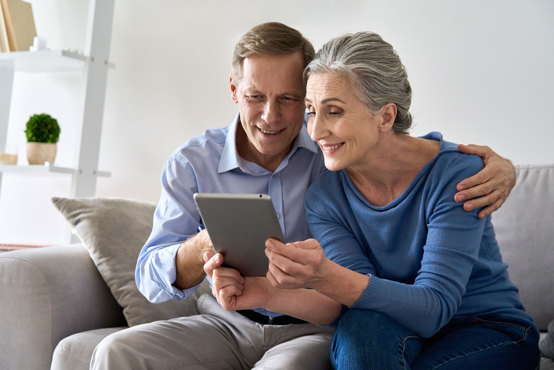 An older couple smiles while looking at a tablet on a sofa