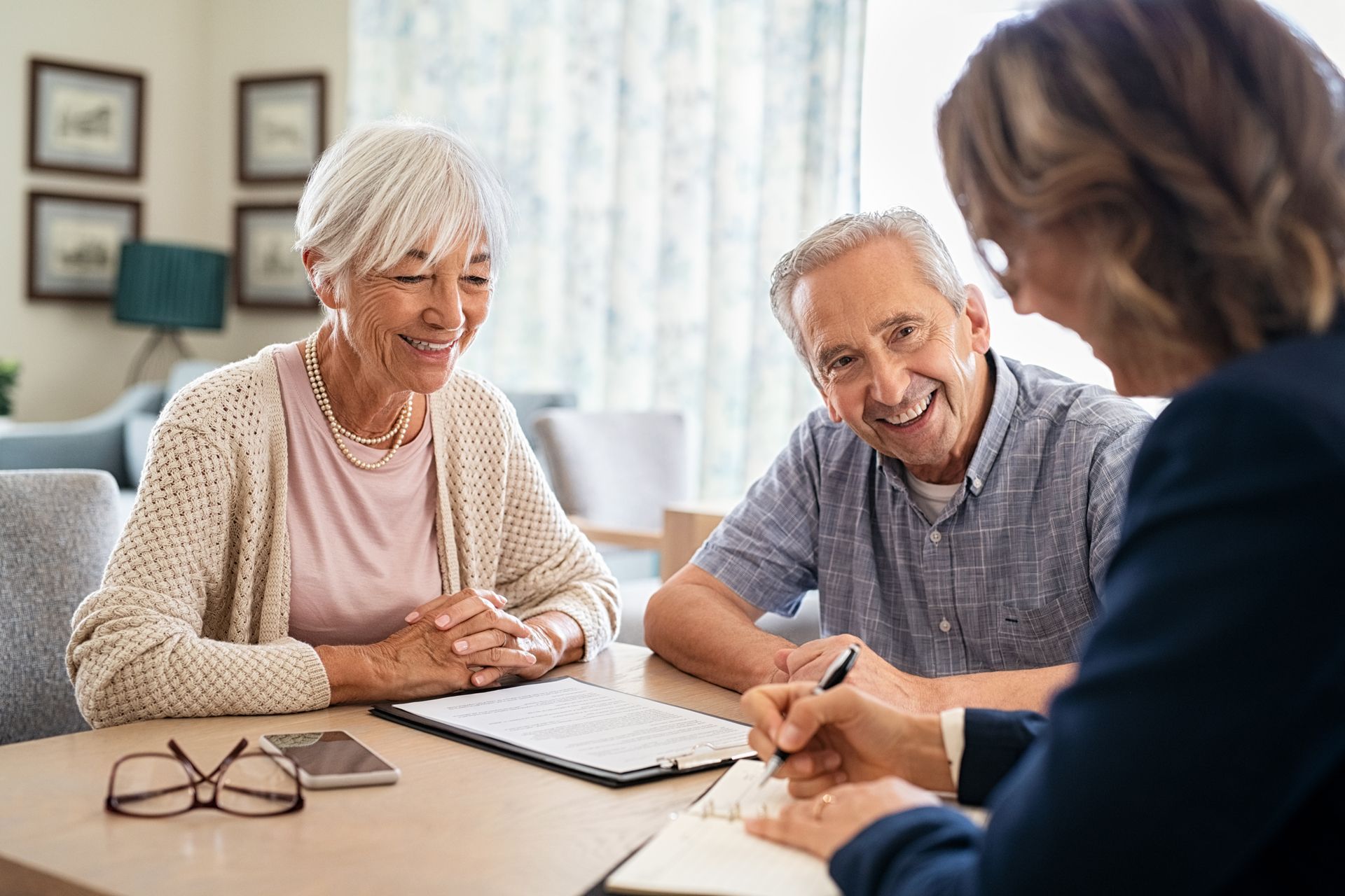 Elderly couple smiling as a person in a suit writes at a table in a home