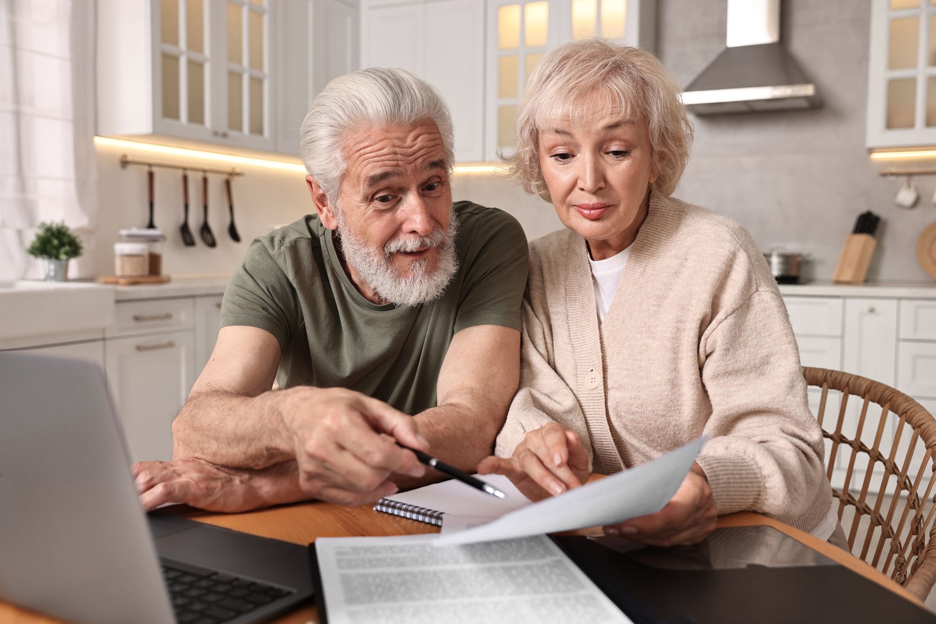 Older couple reviewing paperwork at a table with a laptop in a kitchen