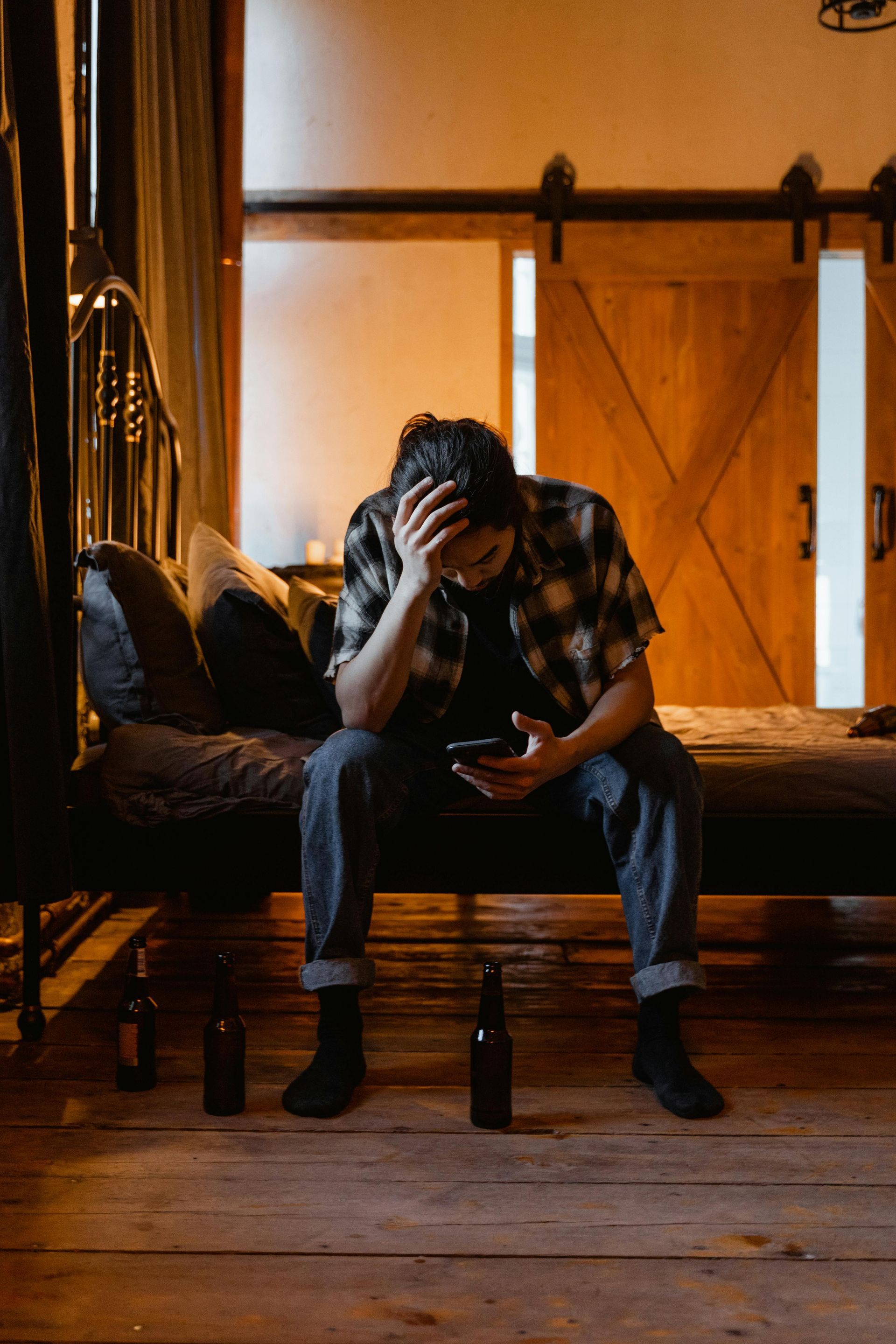 Man with head in hands, sitting on bed, looking at phone; beer bottles on floor.