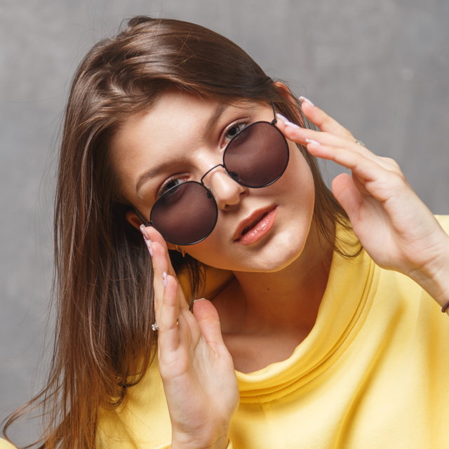 A person in a yellow top adjusts dark-rimmed sunglasses, looking forward against a neutral gray background.