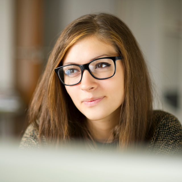 A person with shoulder-length brown hair and glasses looking thoughtfully to the side against a blurred background.