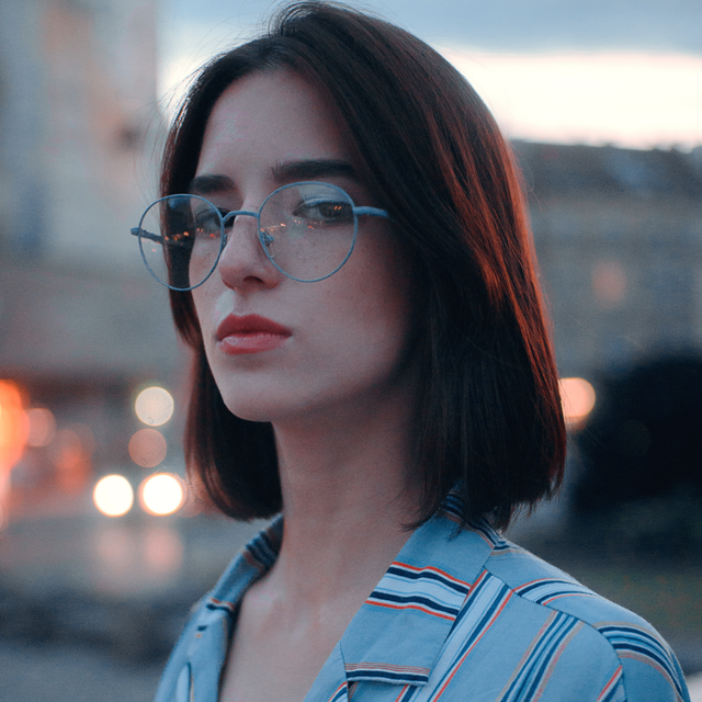 A person with short dark hair wears round, light-blue glasses and a striped blue shirt against a soft, city-light bokeh.