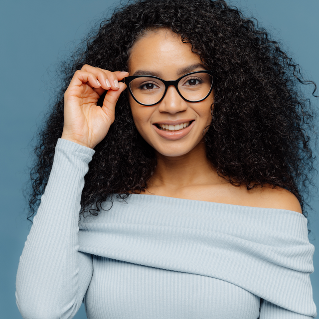 A person with curly dark hair wearing glasses and a light blue off-the-shoulder top, smiling against a blue background.