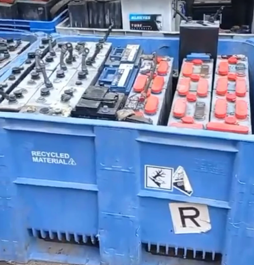 Contenitore blu pieno di batterie usate da riciclare; etichette di pericolo visibili.