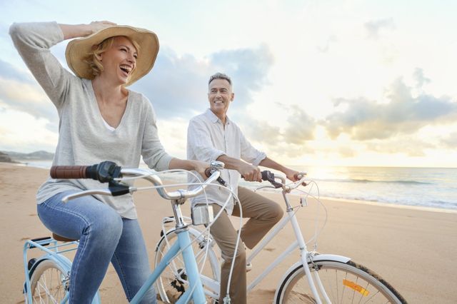 Two people ride bicycles along a sunny beach, smiling near the shoreline.