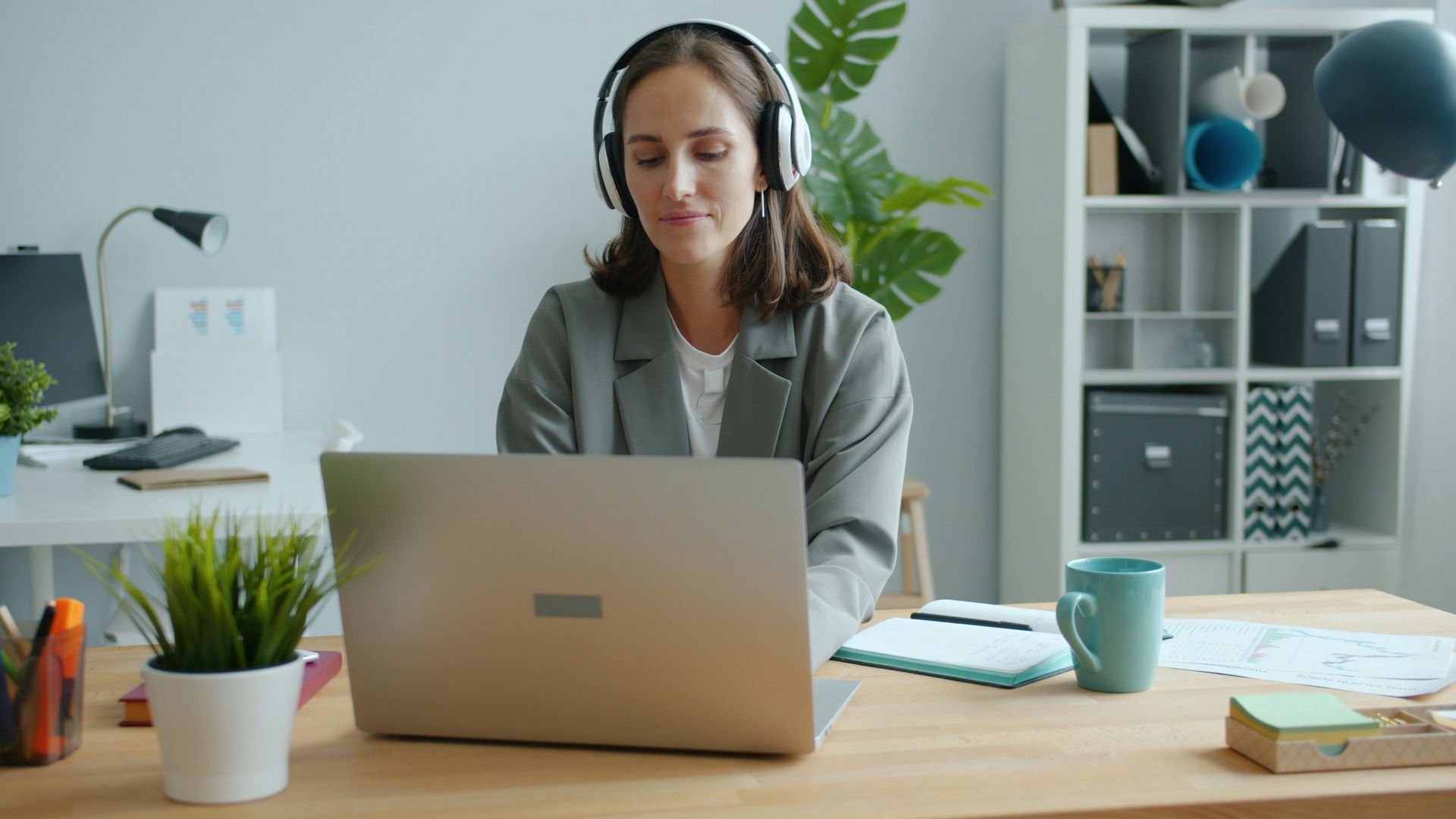 Woman wearing headphones, working on a laptop at a desk. Office setting, with potted plants, and other office supplies.