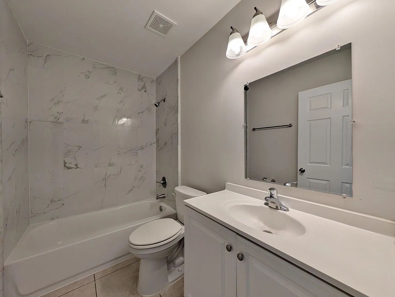 Bathroom with white fixtures: tub, toilet, and vanity with a mirror and overhead light. White marble-look shower surround.