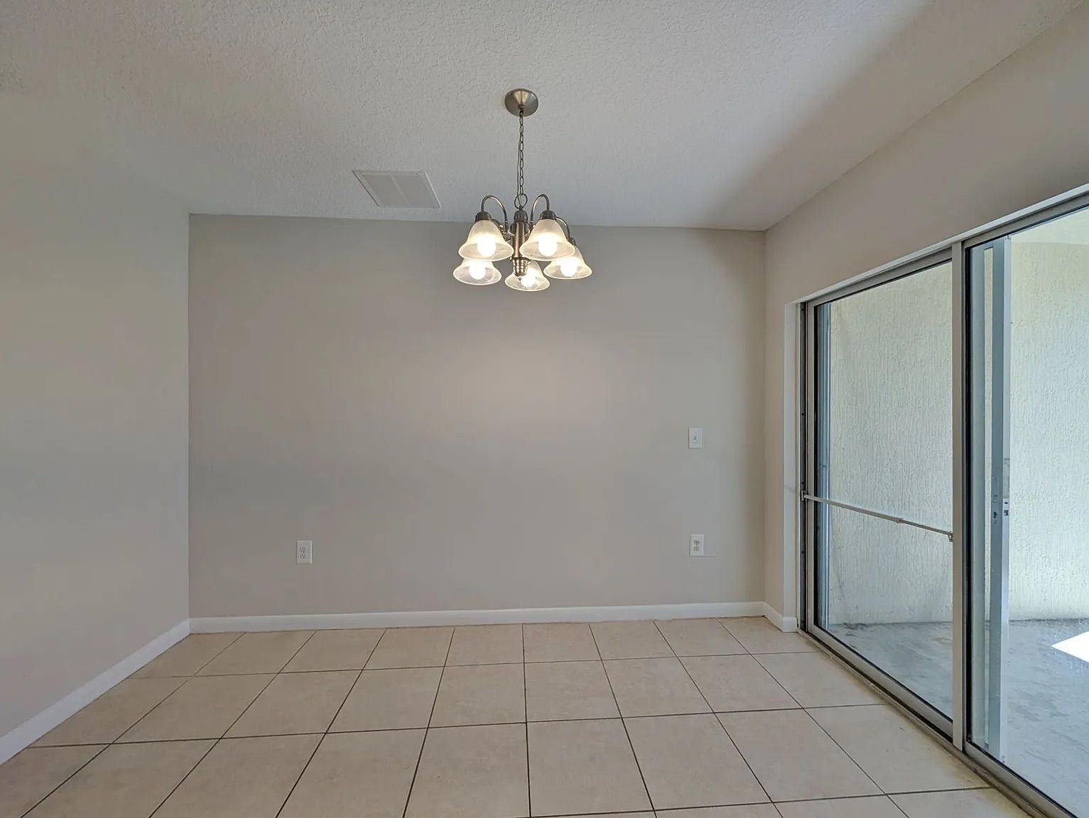 Empty dining room with beige tiled floor, neutral walls, and a chandelier. Sliding glass door visible.