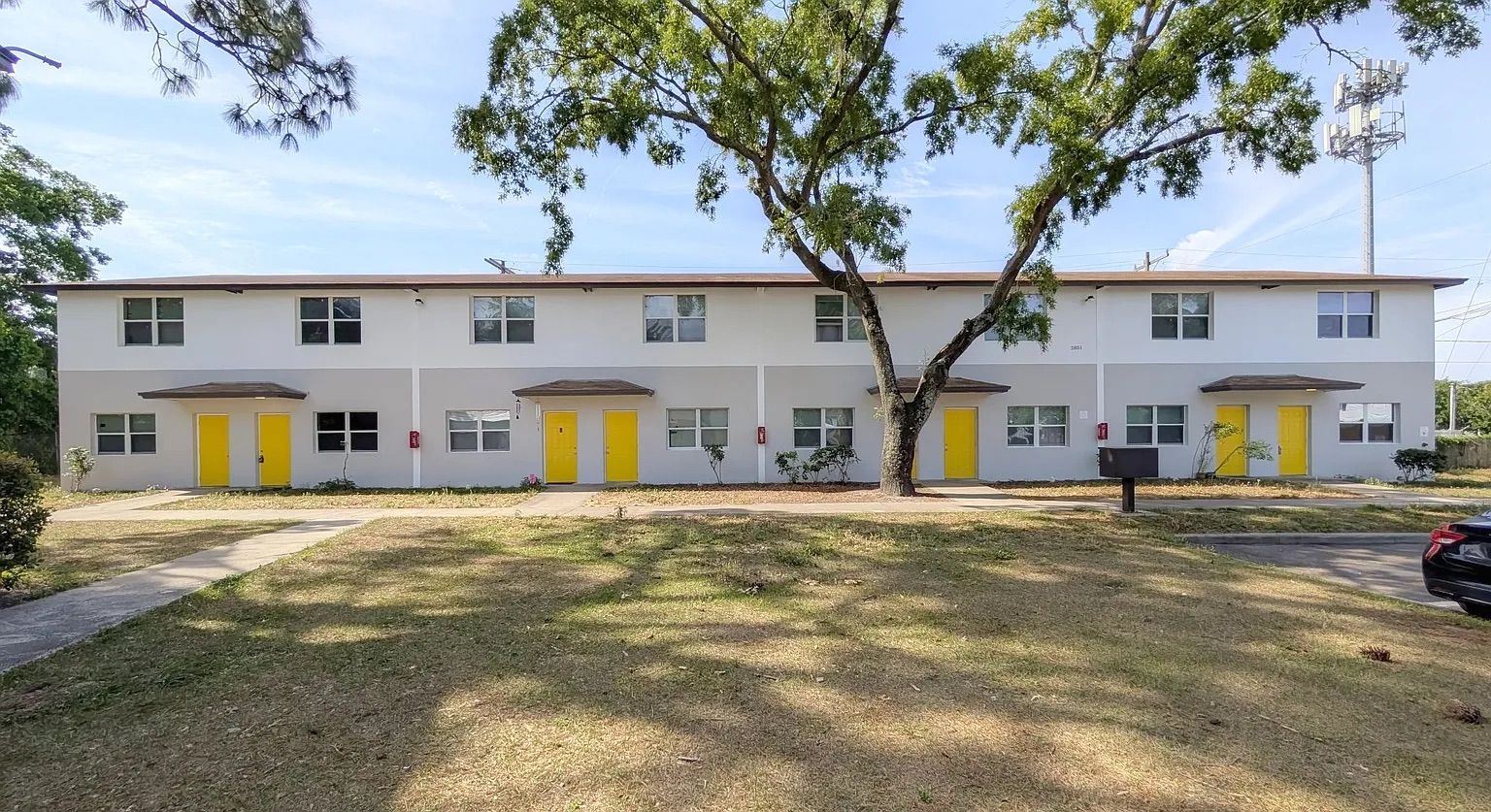 Two-story apartment building with yellow doors, gray walls, and a tree in front.