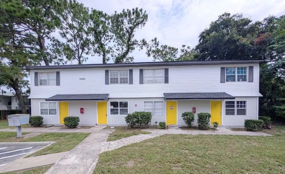 Two-story apartment building with yellow doors and gray siding, set against a backdrop of trees and a cloudy sky.
