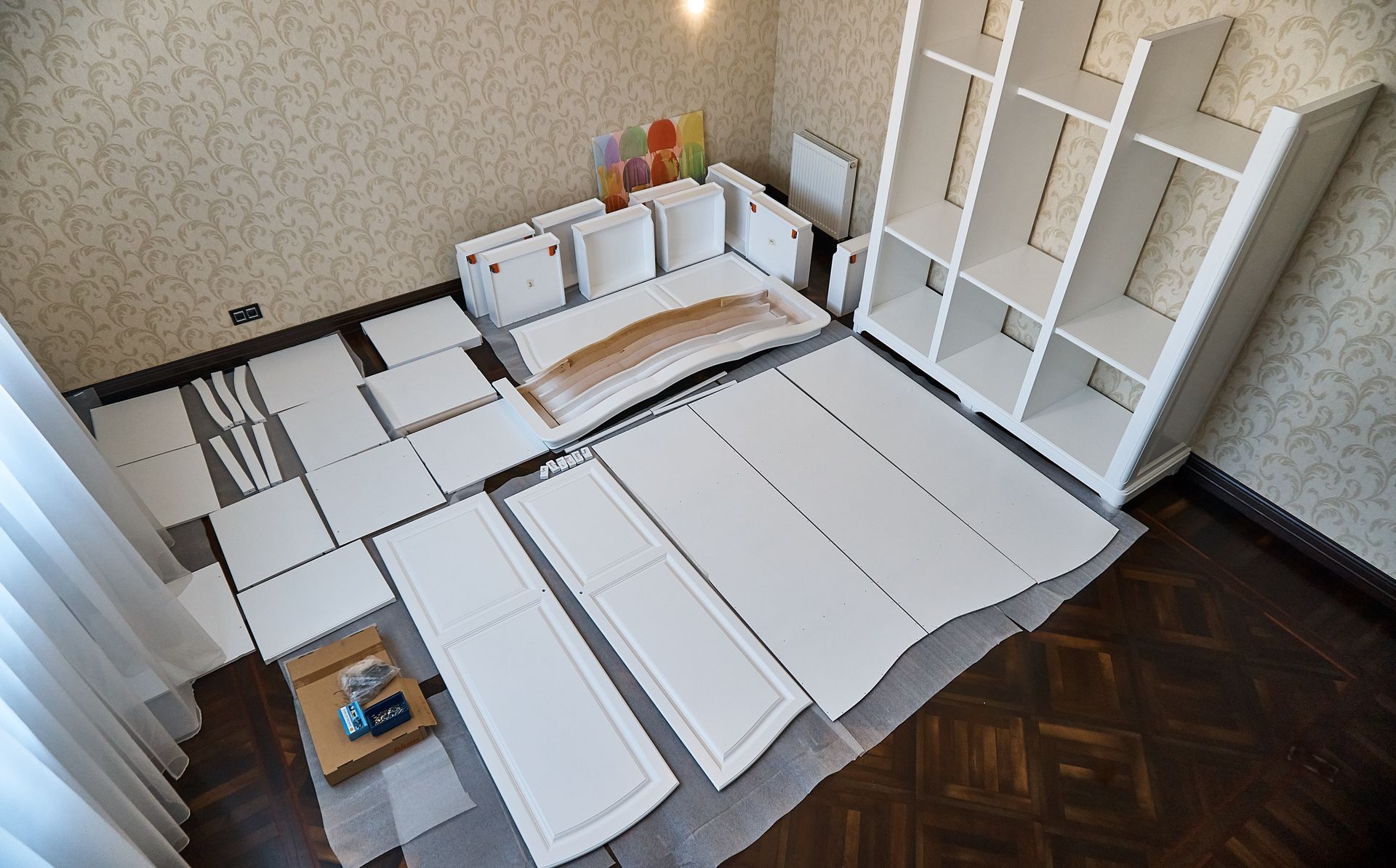 Unassembled white shelving unit parts and assembled bookcase on a floor with parquet and wallpaper.