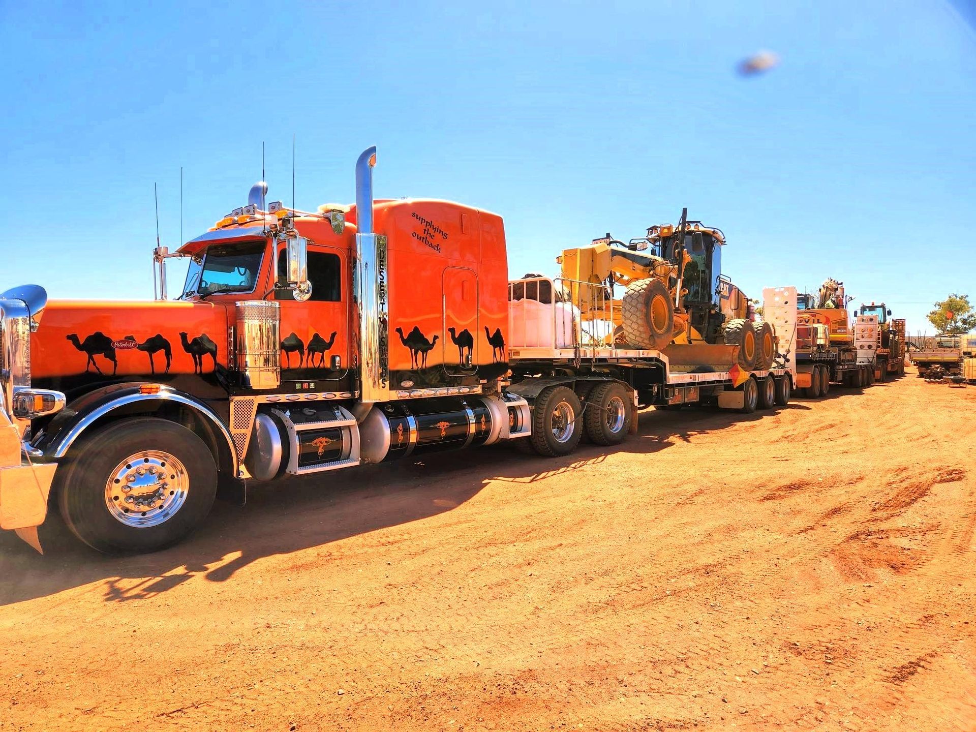 A red semi truck with camels painted on the side is parked in a dirt field.