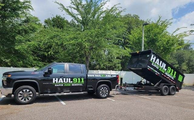 A black Haul 911 pickup truck towing an elevated dump trailer in an outdoor parking lot with trees in the background.