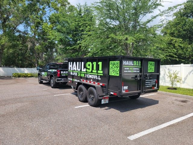 A black pickup truck towing a large black junk removal trailer parked in an outdoor lot on a sunny day.
