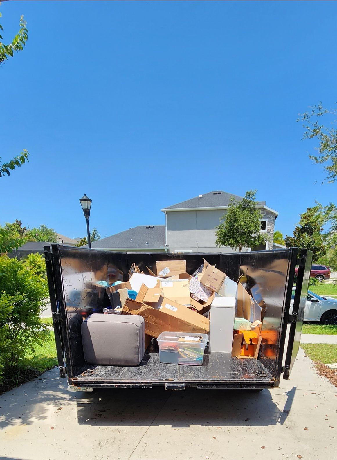 A dumpster filled with assorted trash and cardboard boxes sits on a suburban driveway under a clear blue sky.