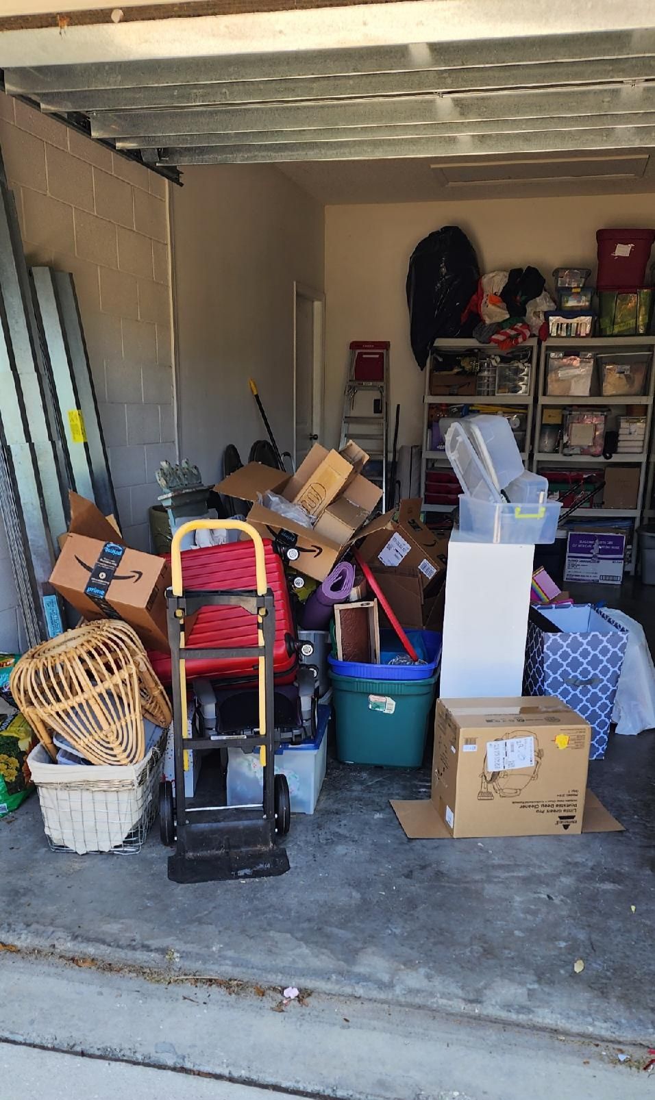 A cluttered garage interior filled with cardboard boxes, storage bins, a hand truck, and a wicker chair on a concrete floor.