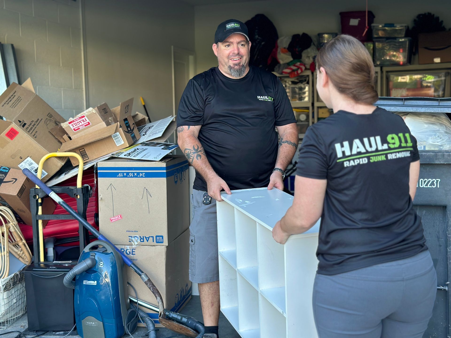 Two people in Haul 911 shirts carry a white cube organizer from a garage filled with boxes and equipment.