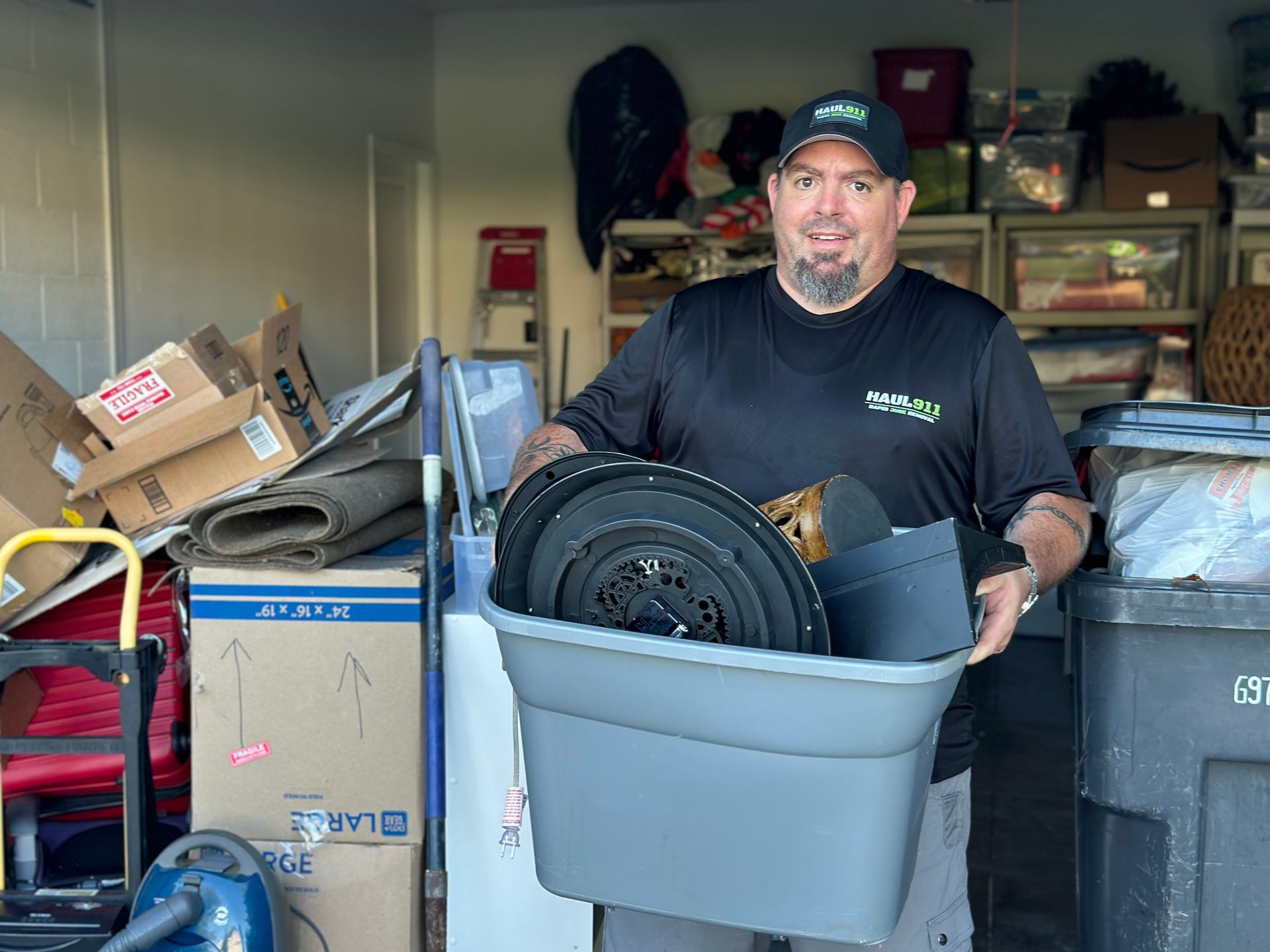A person wearing a black shirt and cap smiles while holding a grey storage bin in a garage filled with boxes and items.