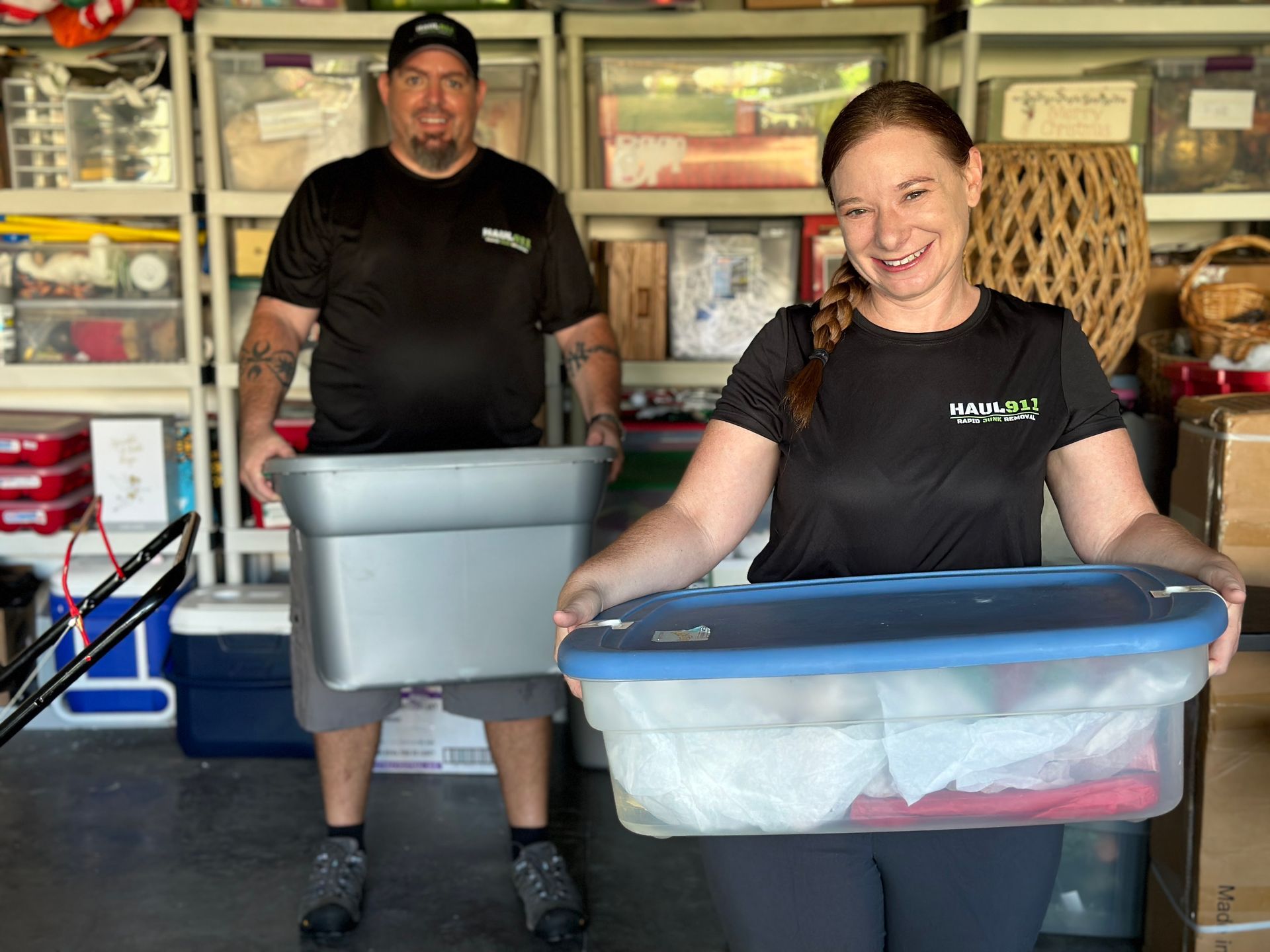 Two smiling people wearing matching black shirts hold large plastic storage bins in a cluttered garage.