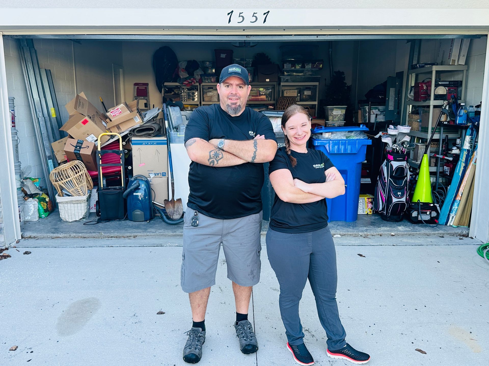 A man and woman with arms crossed stand in front of a garage packed with boxes and miscellaneous items.