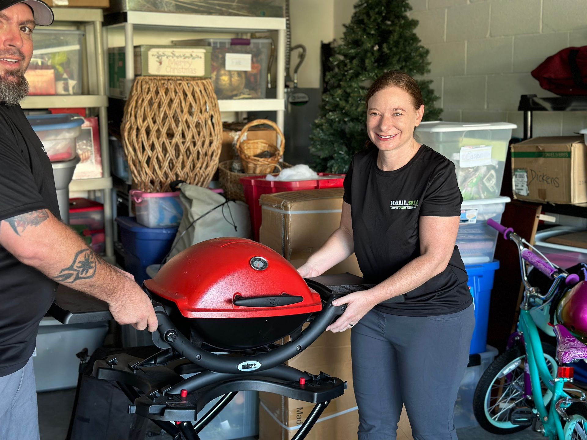 Two people stand in a storage unit holding a portable red grill between them, surrounded by boxes and household items.