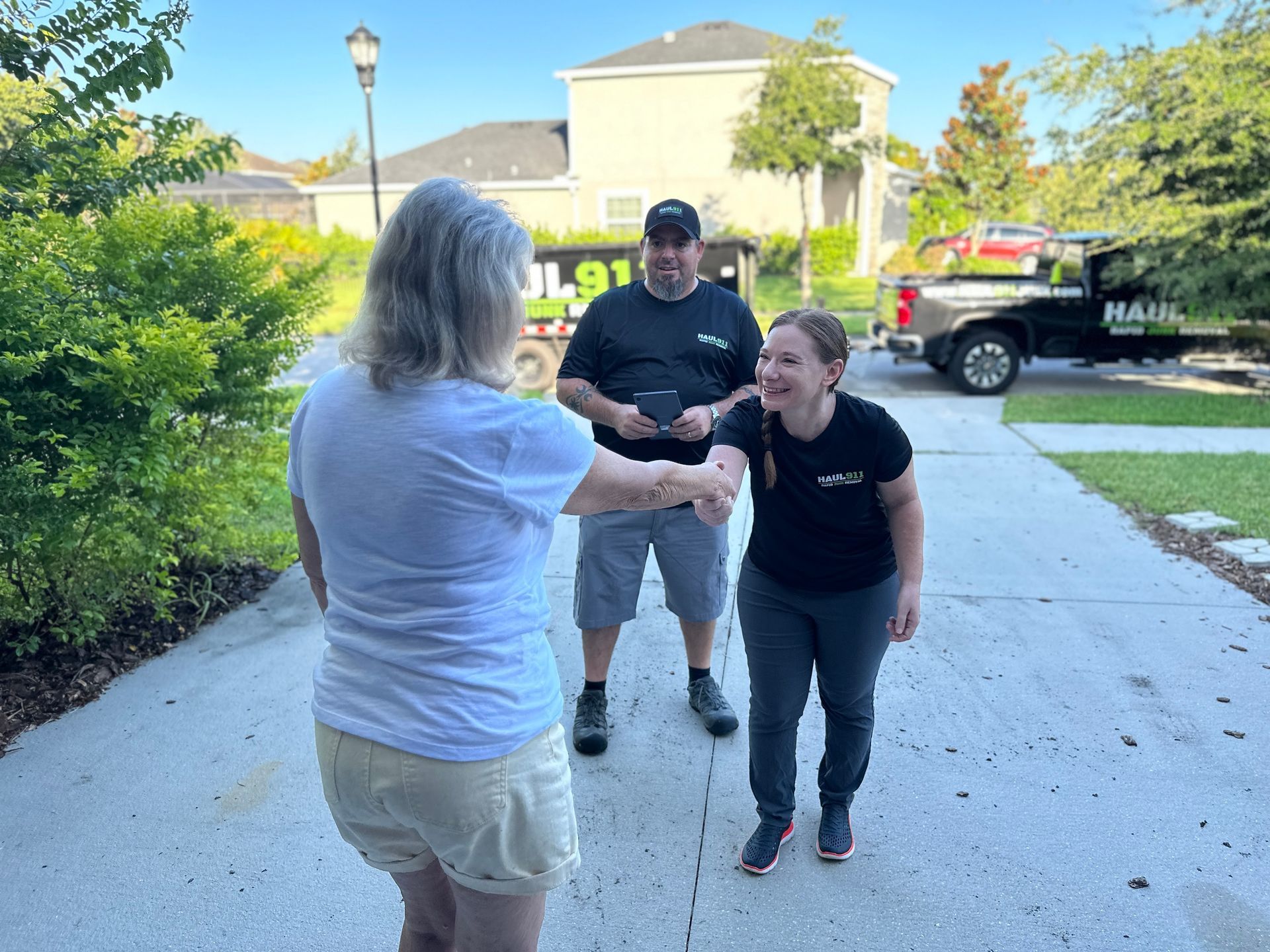 Two people shaking hands on a driveway, with a person standing behind them and a utility truck in the background.