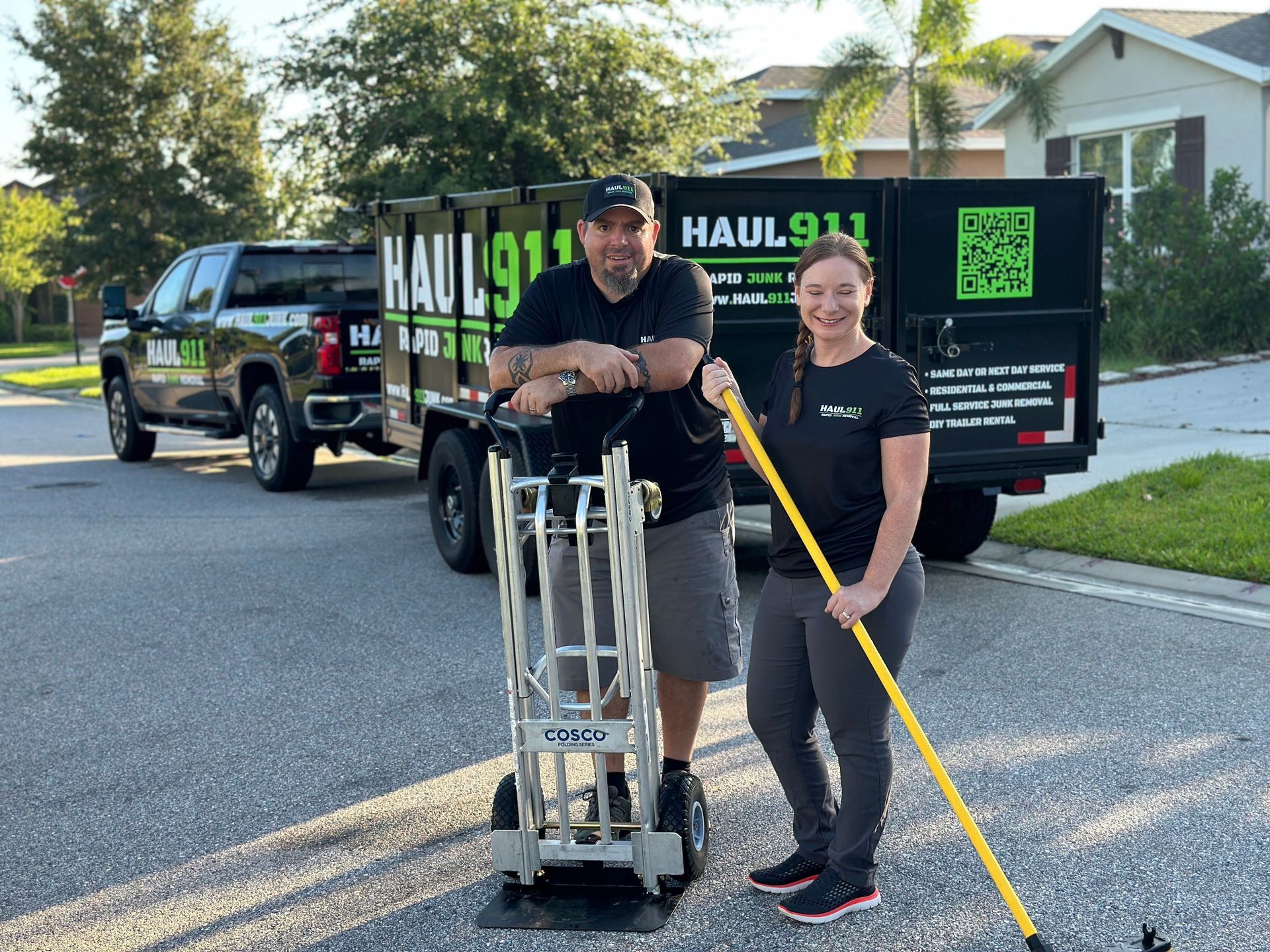 Two people stand in a residential street in front of a Haul 911 truck and dump trailer, holding a hand truck and a rake.