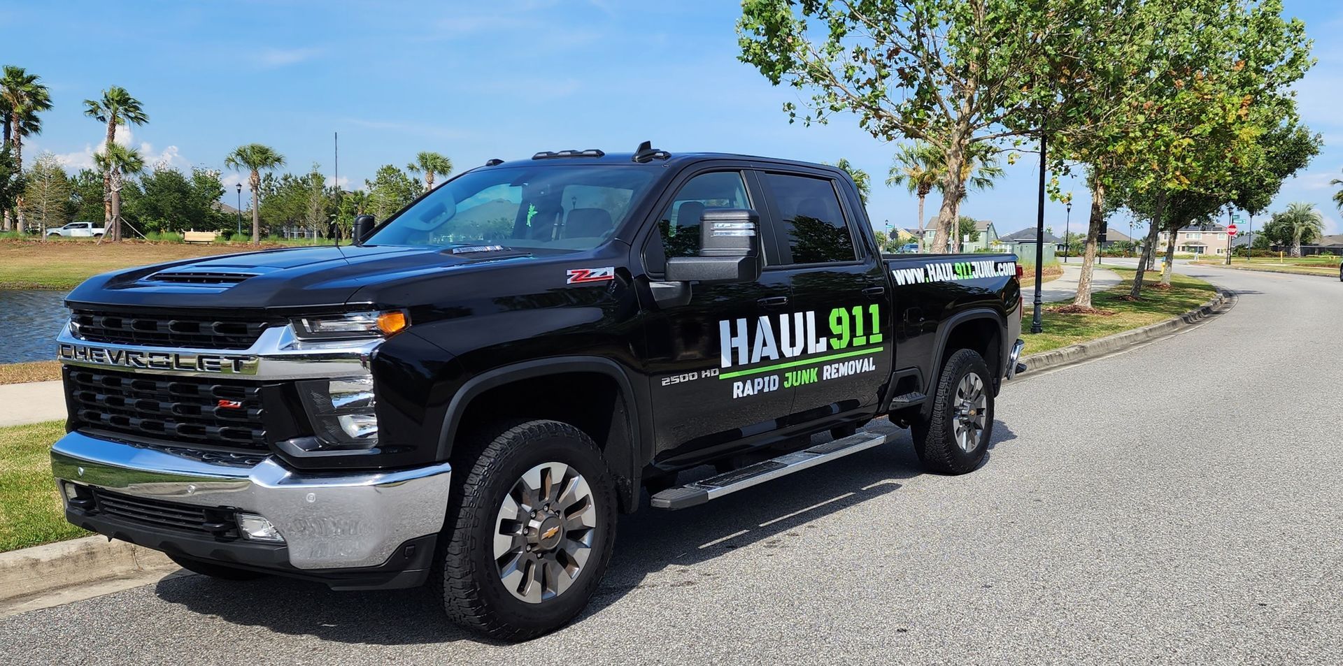 A black Haul911 pickup truck parked on a gravel path near palm trees and a pond.