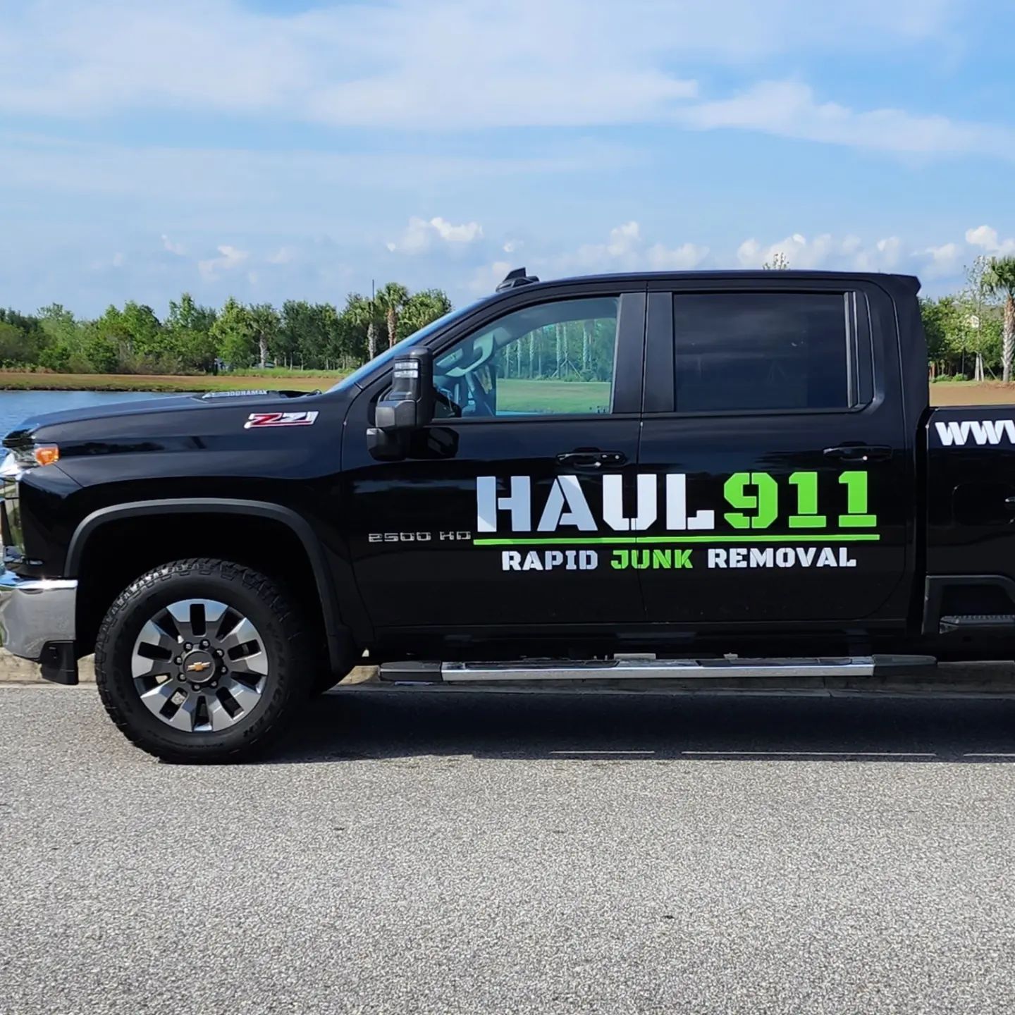 A black Haul 911 junk removal truck parked outdoors against a backdrop of trees and a pond.