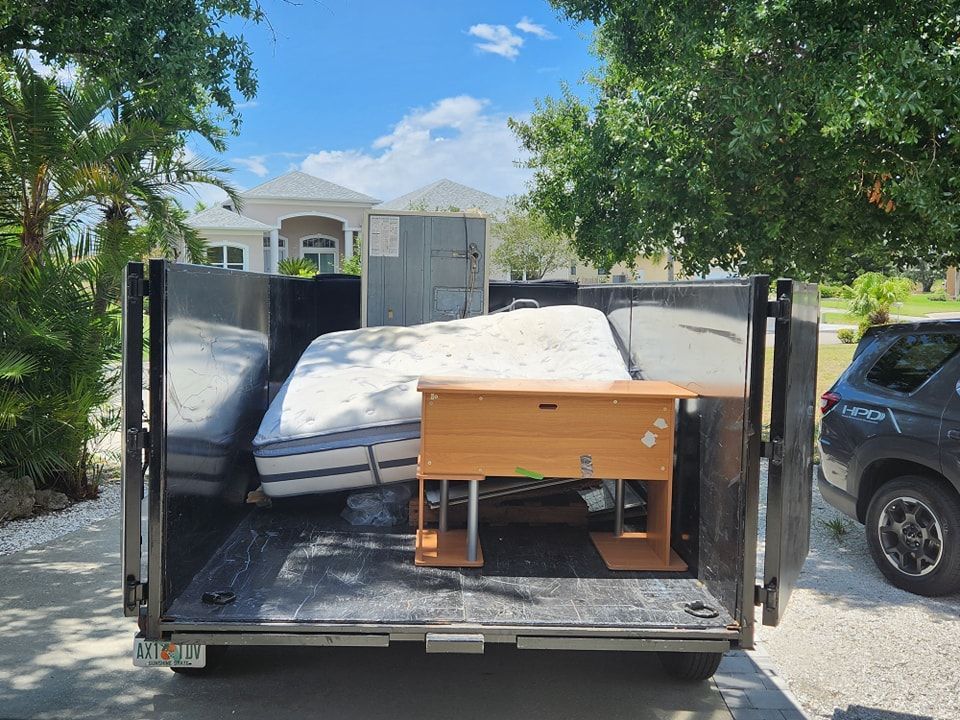 A trailer parked in a driveway holds a mattress, a wooden desk, and a gray metal cabinet, viewed from the rear.