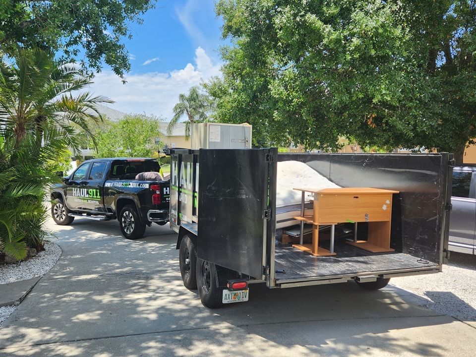 A black pickup truck parked in a driveway towing a trailer filled with furniture and a mattress.