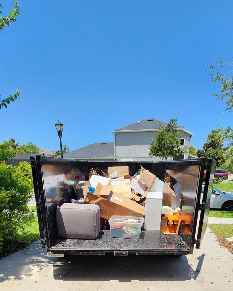 A metal dumpster sits in a driveway, partially filled with cardboard boxes, a grey cushion, and a plastic bin.