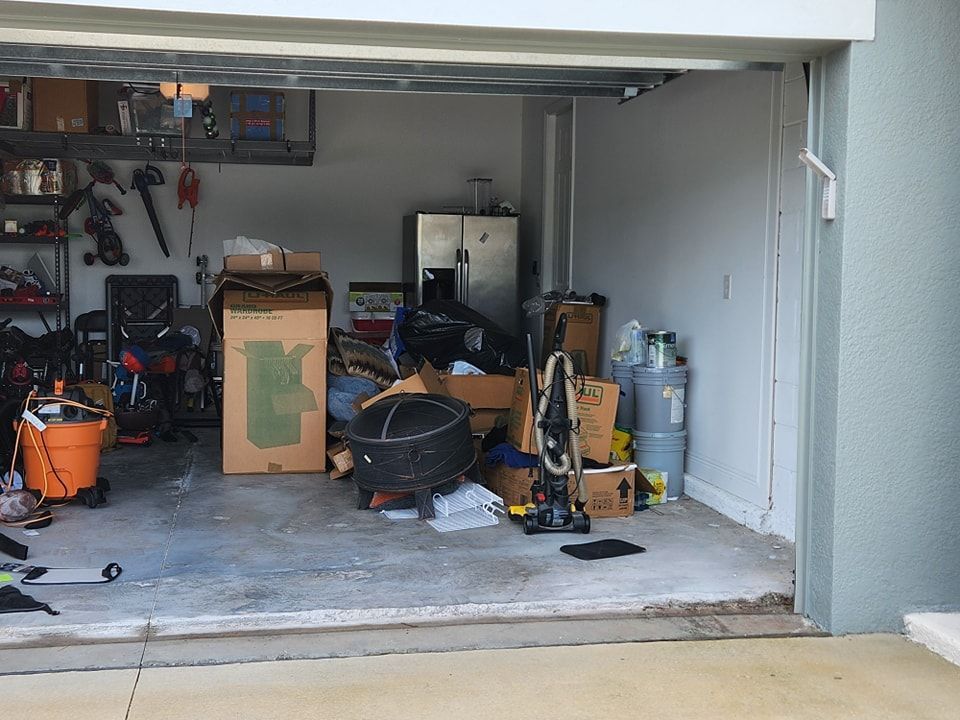 An open, cluttered residential garage filled with cardboard boxes, a refrigerator, and various household items.