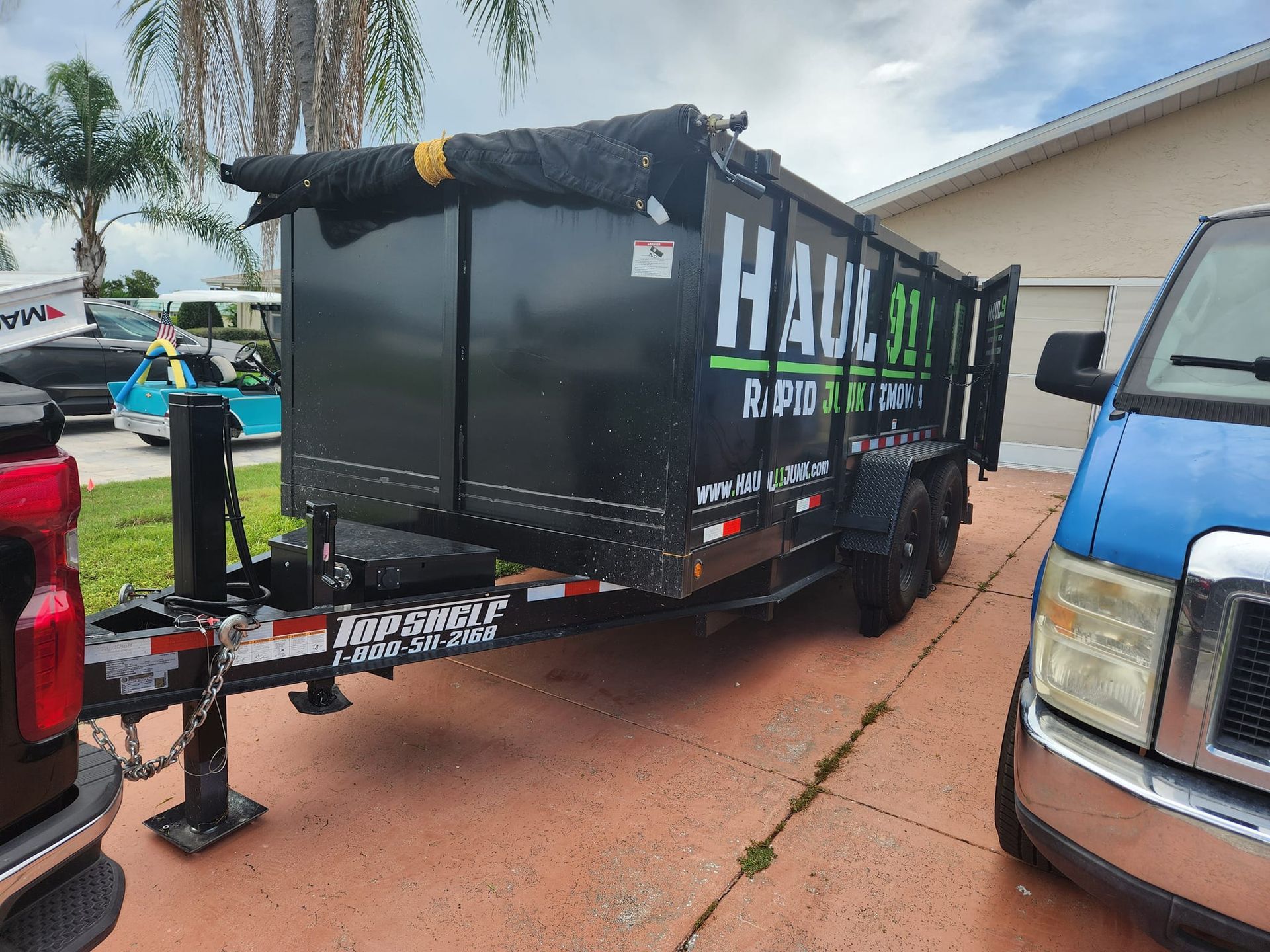 A black metal dump trailer for Haul 911 parked on a driveway next to a blue truck.