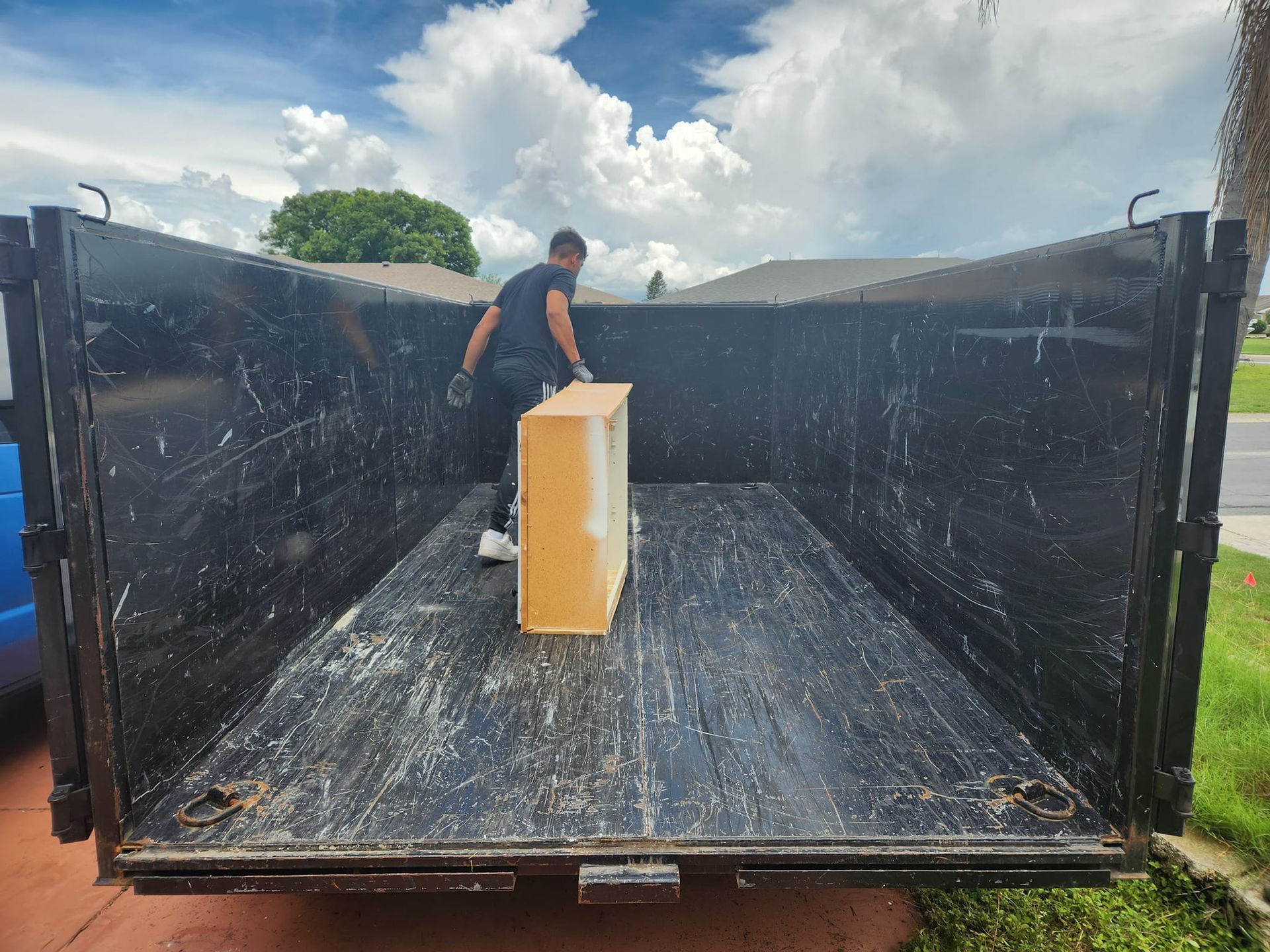 A person stands inside a large, empty black dump trailer on a sunny day, holding a wooden piece of furniture.
