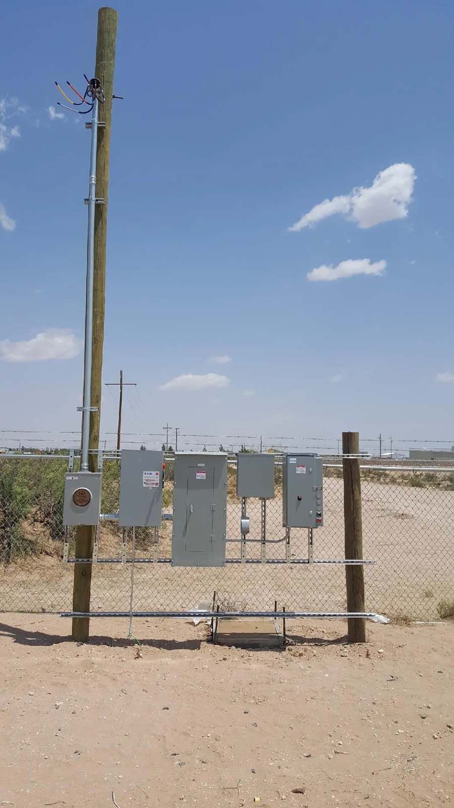 A wooden pole with electrical boxes attached to it is in the middle of a dirt field.