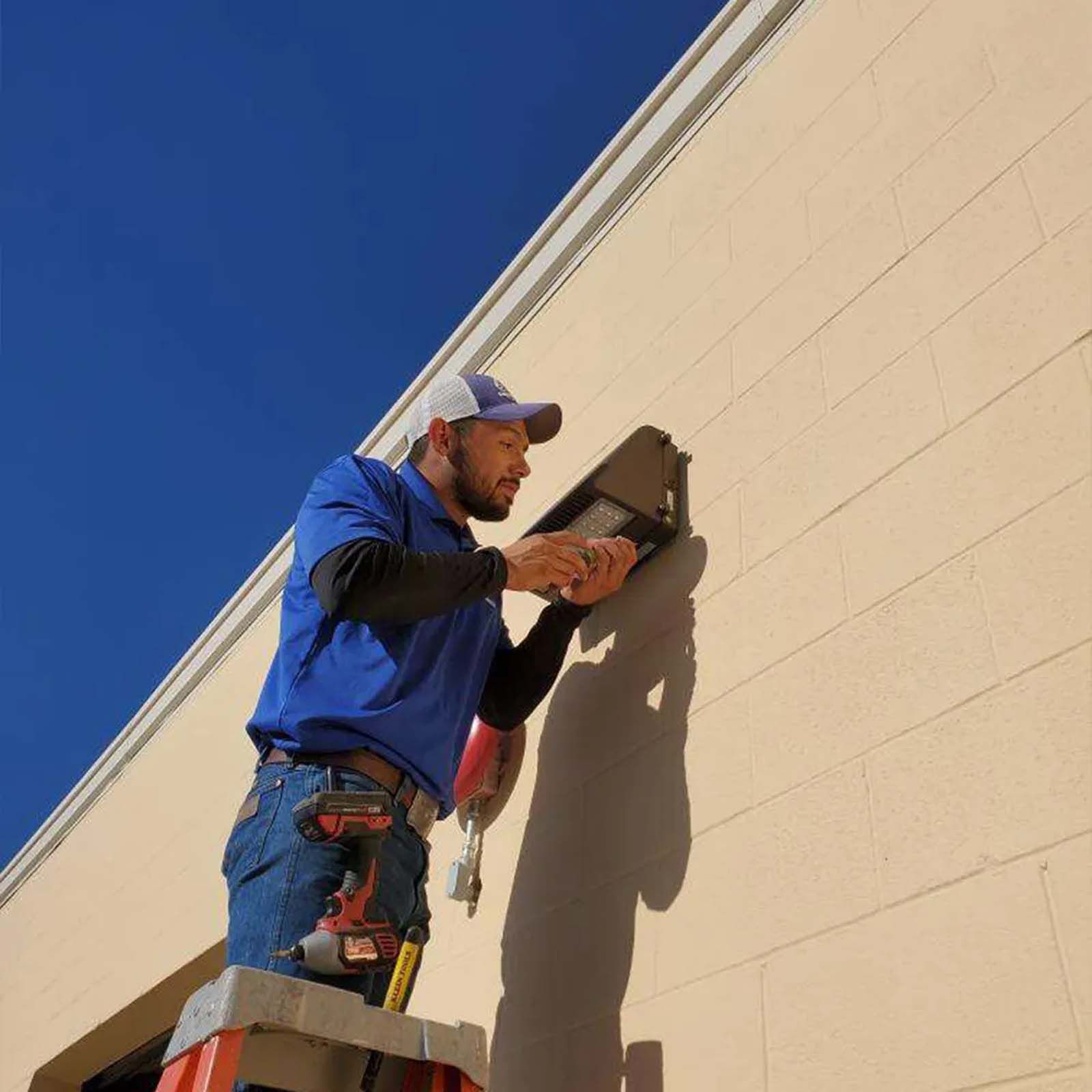 A man is standing on a ladder fixing a light on the side of a building.