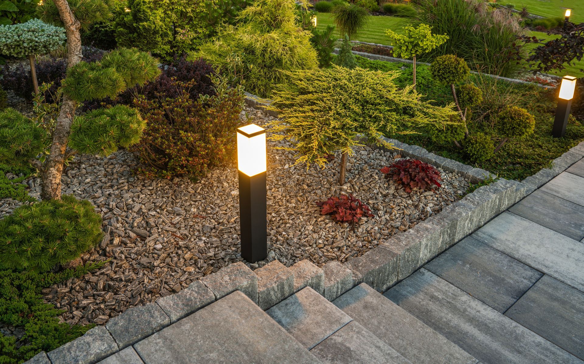 Lit garden path with stone steps, gravel bed, and bushes; pillar lights illuminate the scene at dusk.