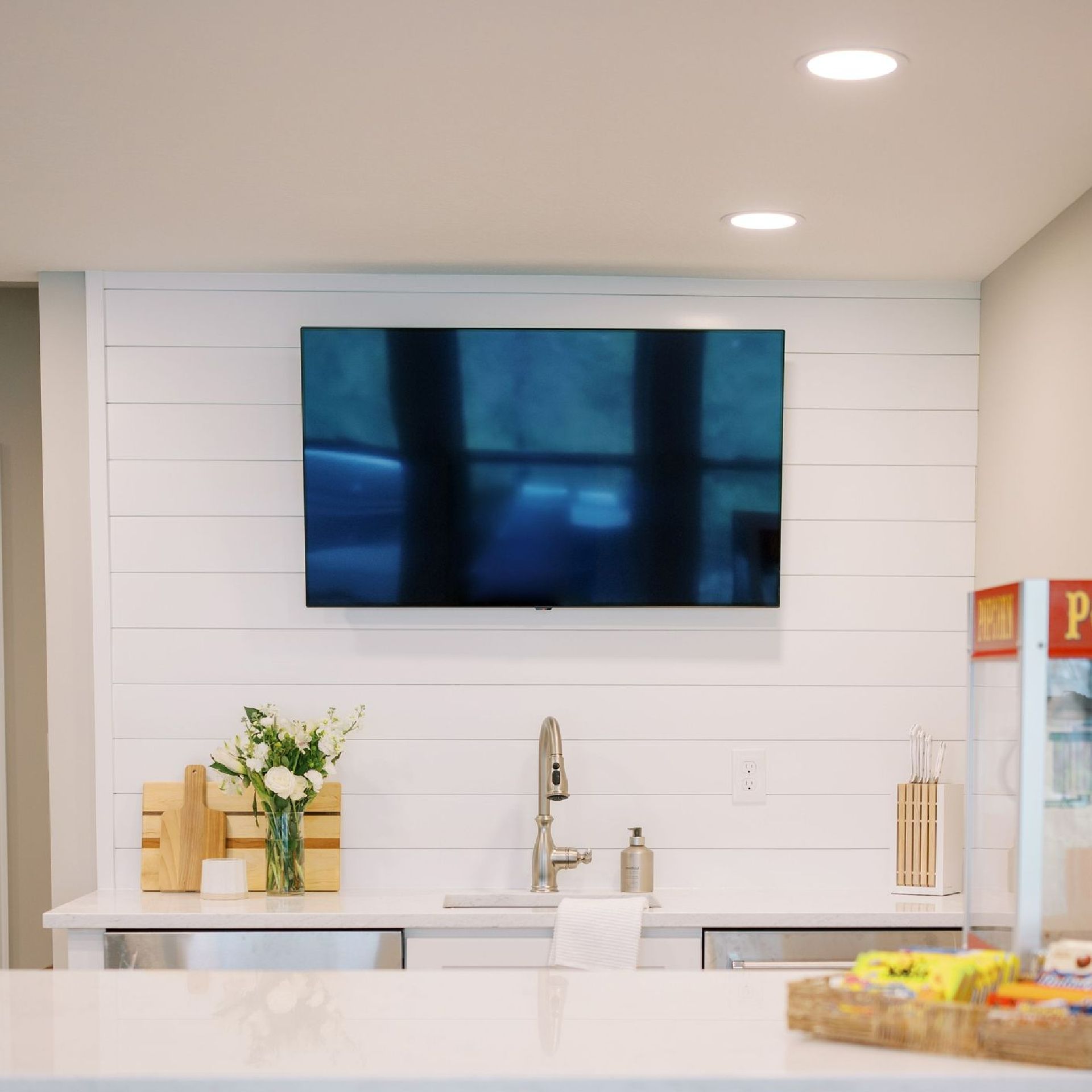 A mounted TV above a bar sink with a white shiplap accent wall. A vase of flowers and a wooden crate sit on the counter