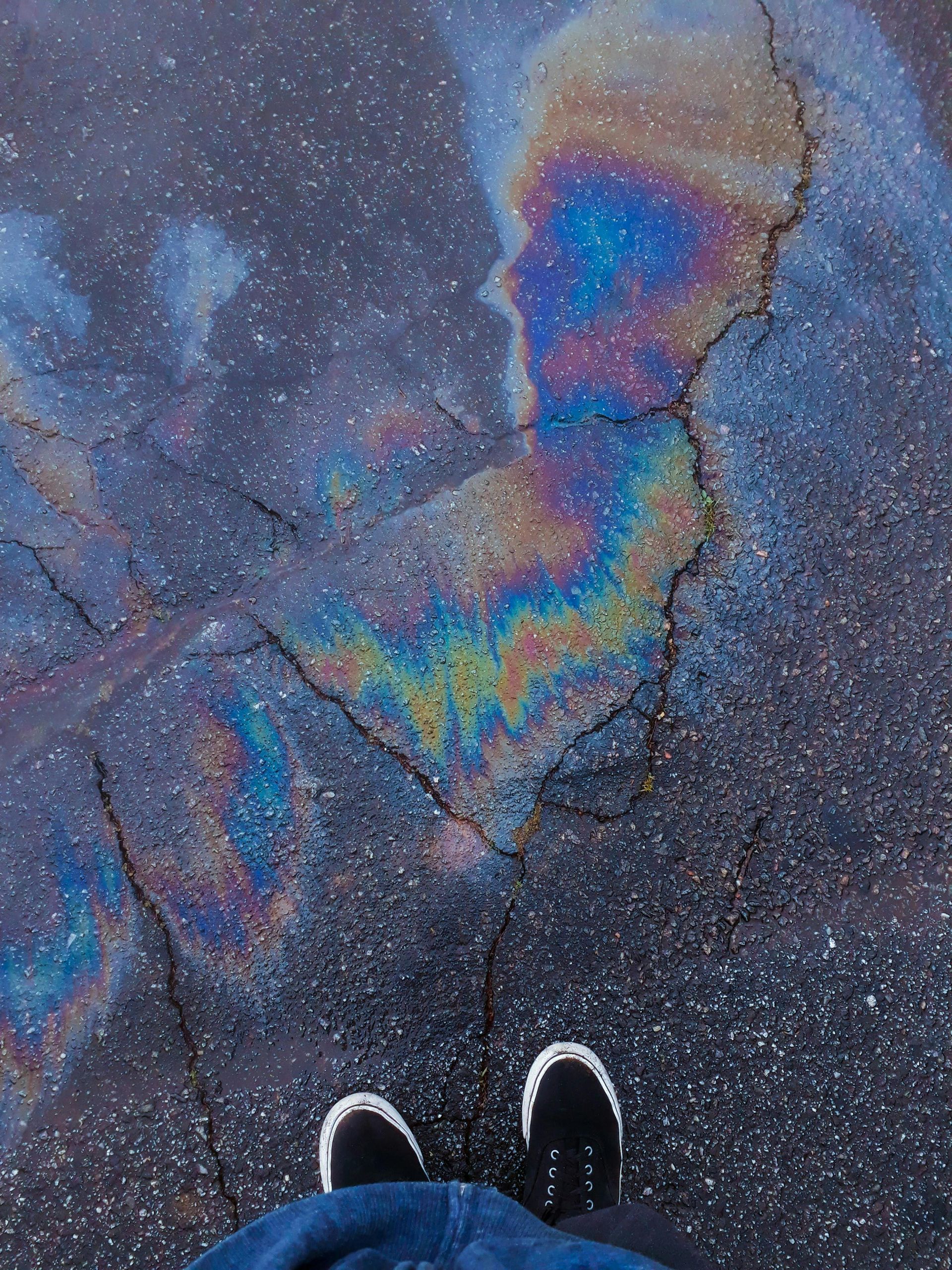 A person is standing in front of a rainbow of oil on the ground.