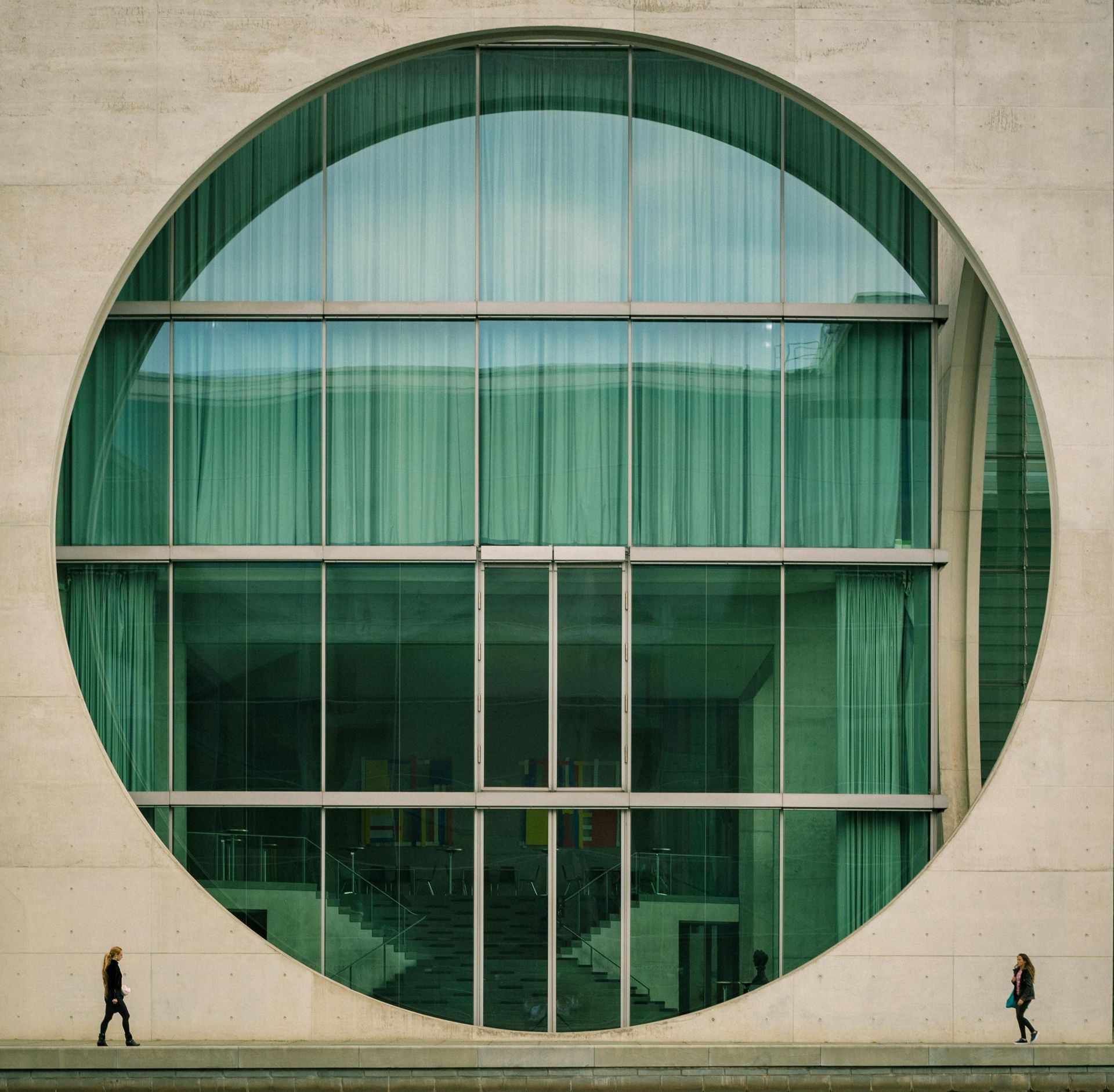 Circular window on a modern building, framed by concrete, with two figures walking outside.