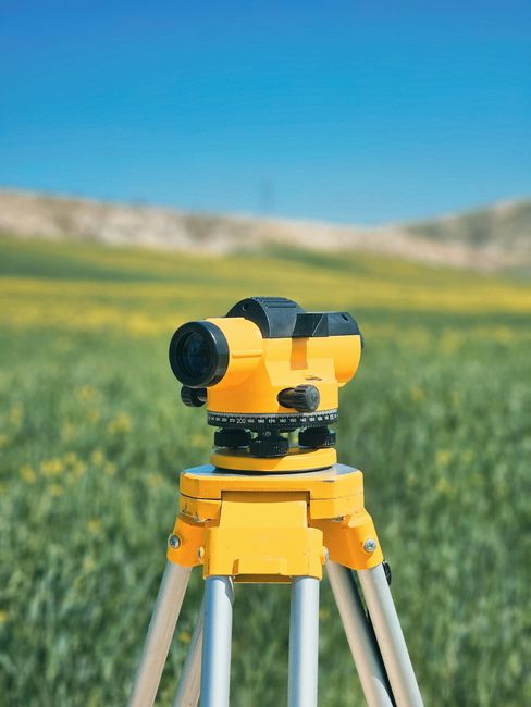 Yellow surveyor's level on a tripod in a field of green plants with a blue sky background.
