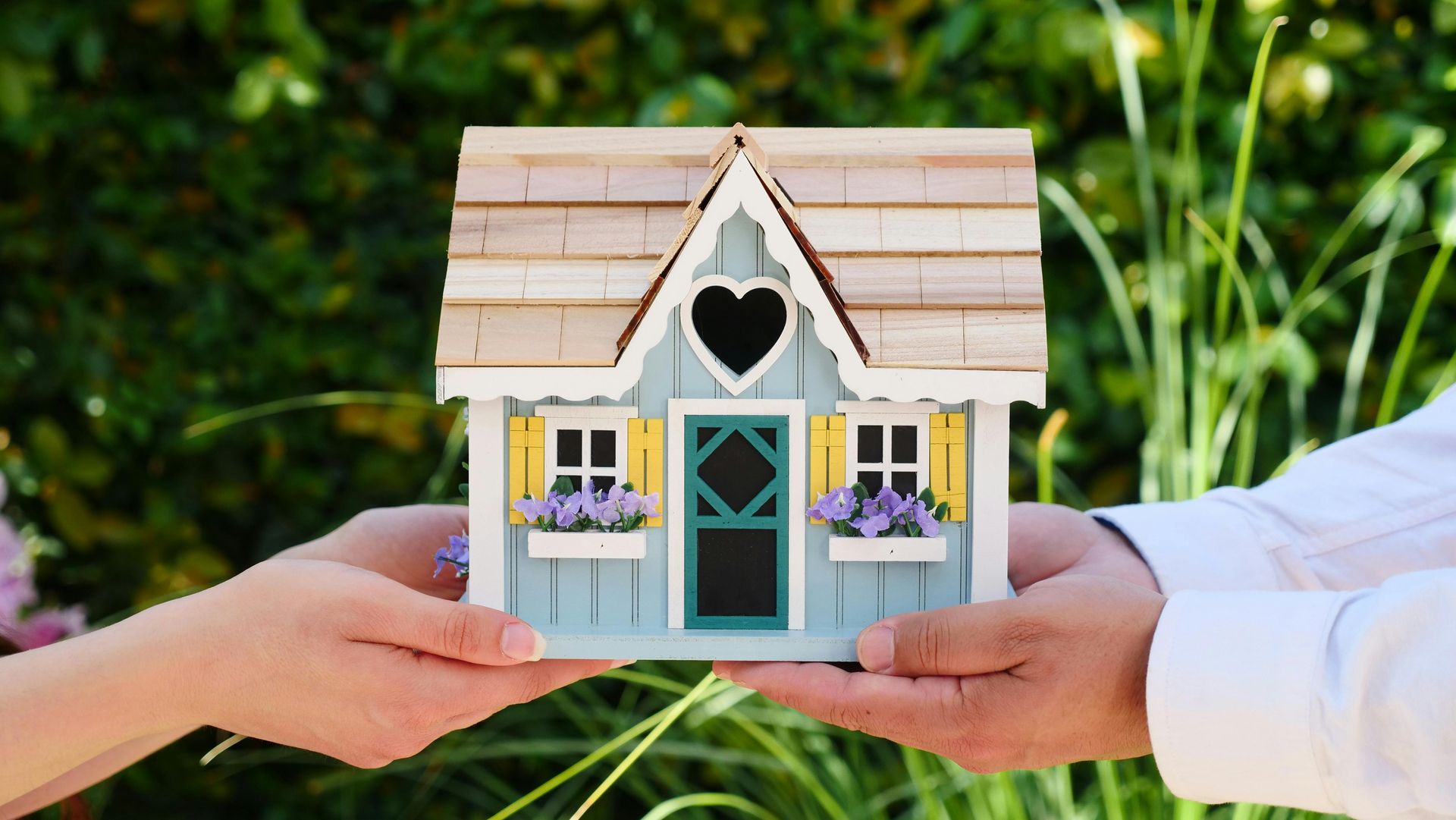 Hands holding a miniature blue house with heart cutout, plants, and yellow shutters.