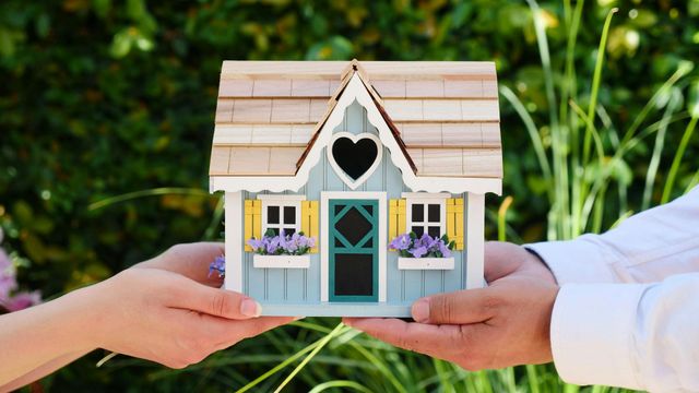 Hands holding a miniature blue house with heart cutout, against a blurred green background.