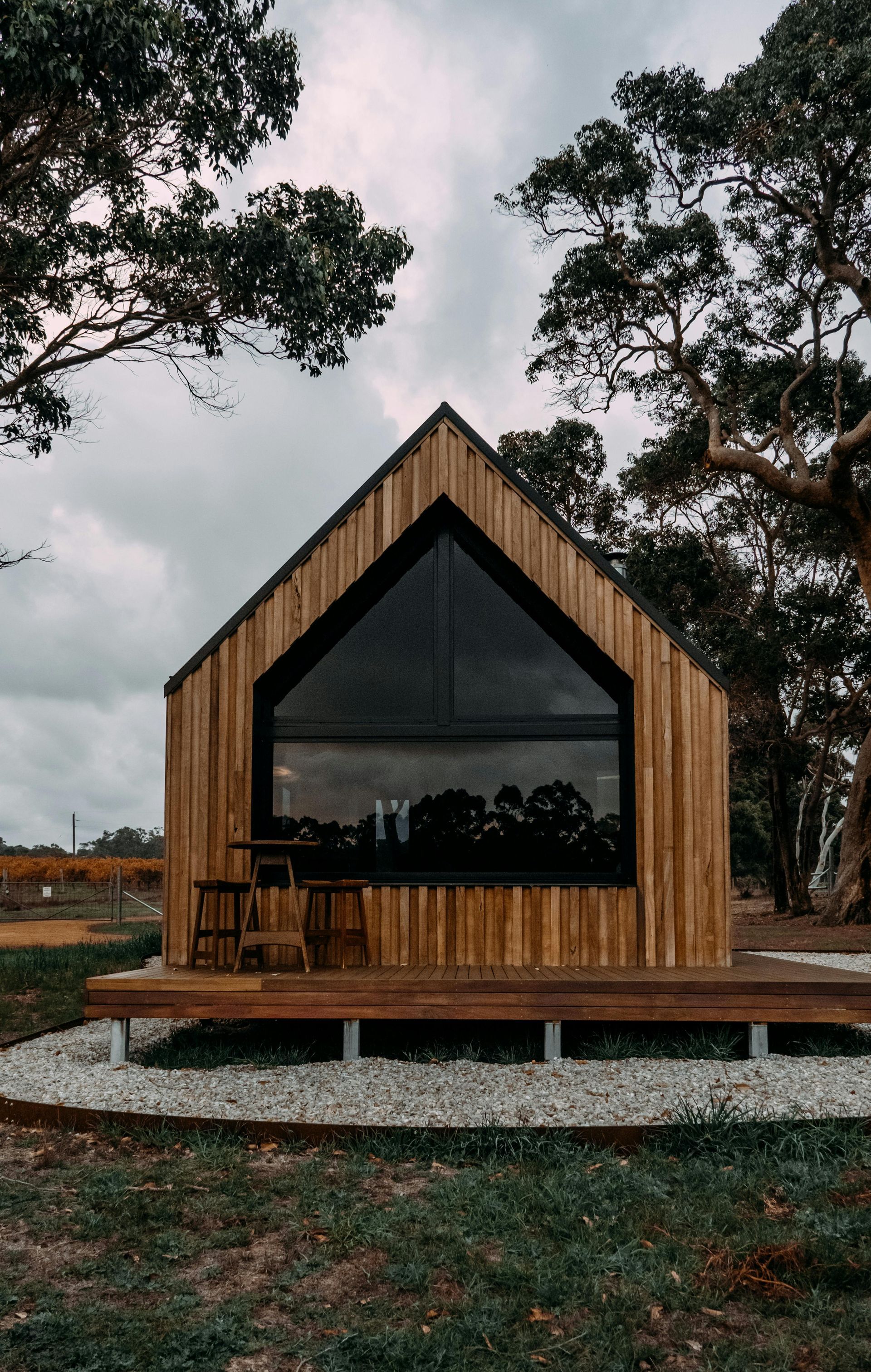 Wooden cabin with a triangular roof and large dark window, set on a raised deck, against a cloudy sky.