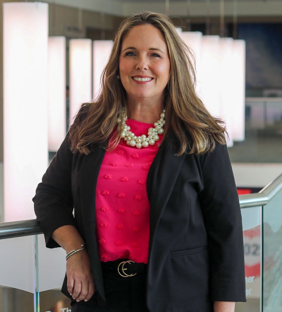 Woman in a black blazer and pink blouse smiling in a modern office hallway.